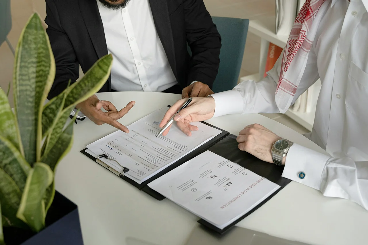 Two men in business suits seated at a table, reviewing documents and discussing details.