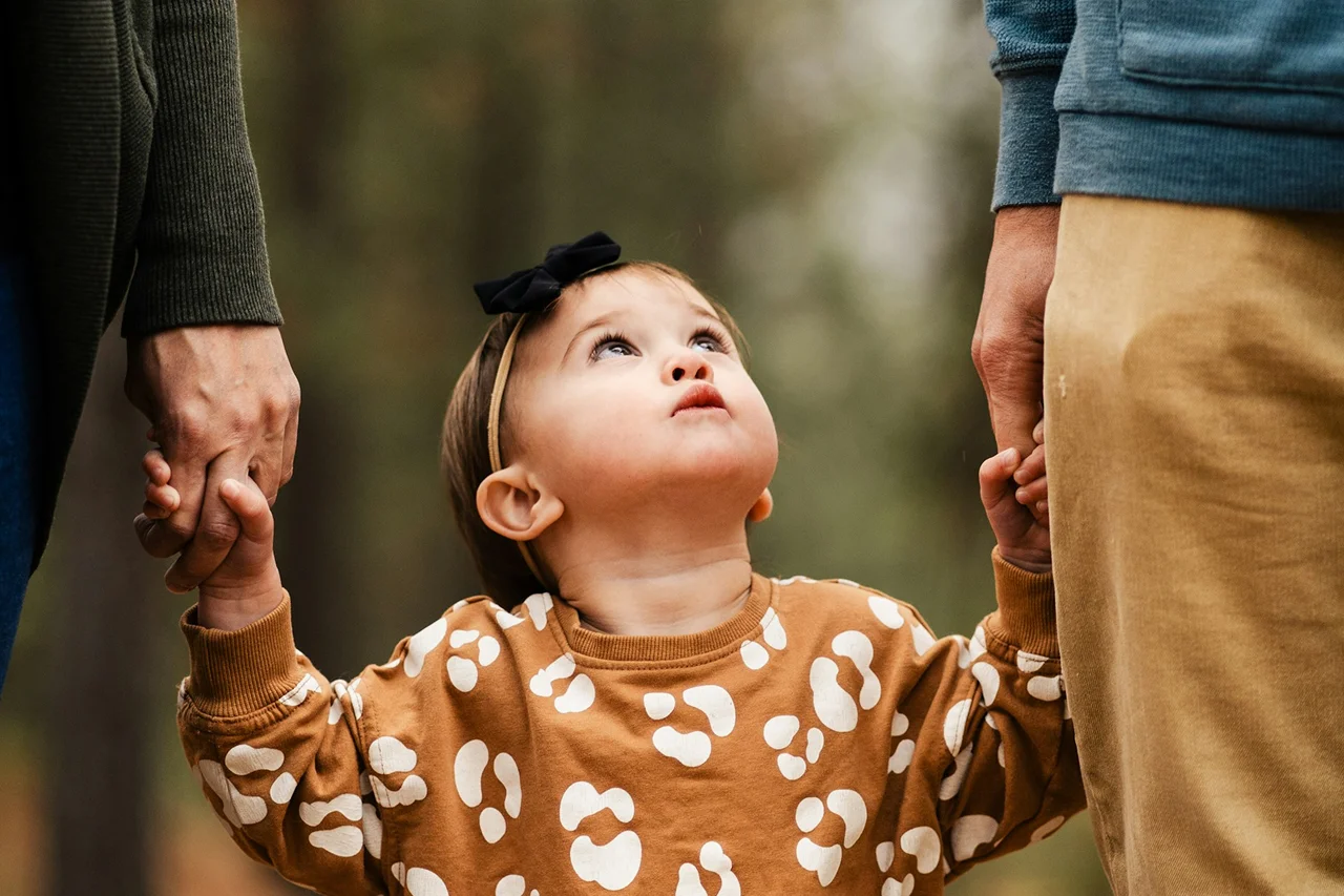 A baby girl gazes up at her parents, who are holding hands, creating a warm family moment.