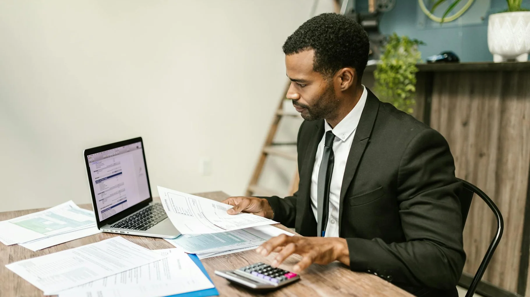 A man in a suit sits at a desk, working on a laptop in a professional office setting.