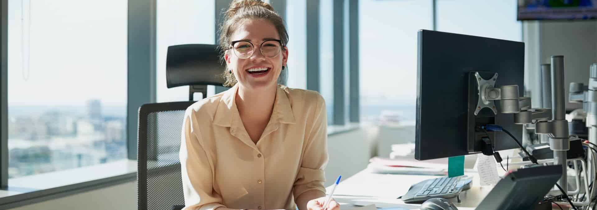 A woman smiles warmly while seated at her desk, exuding a sense of happiness and contentment in her workspace.