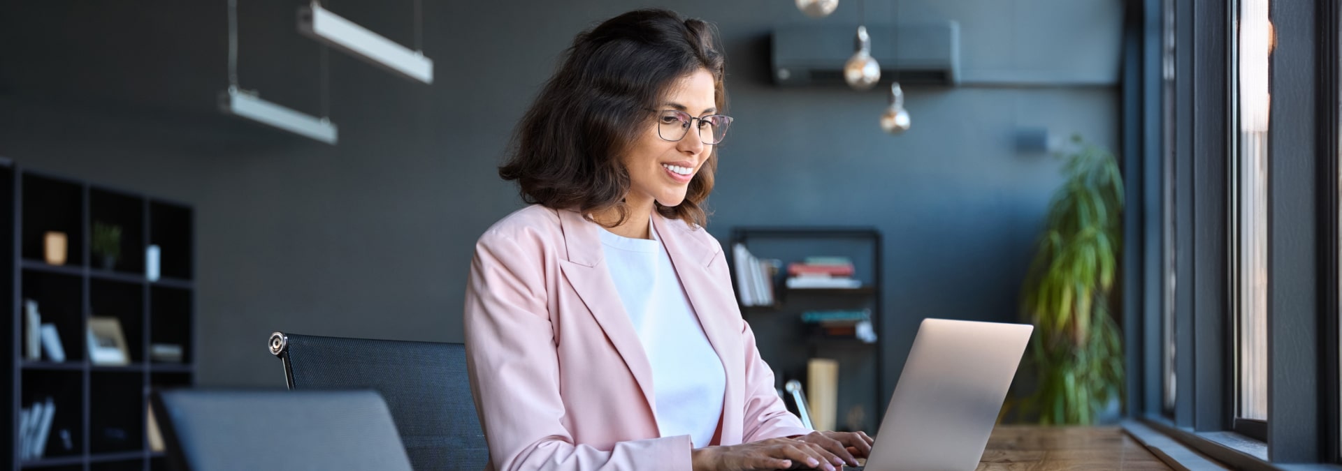A woman wearing glasses sits at a desk, focused on her laptop, surrounded by a tidy workspace.
