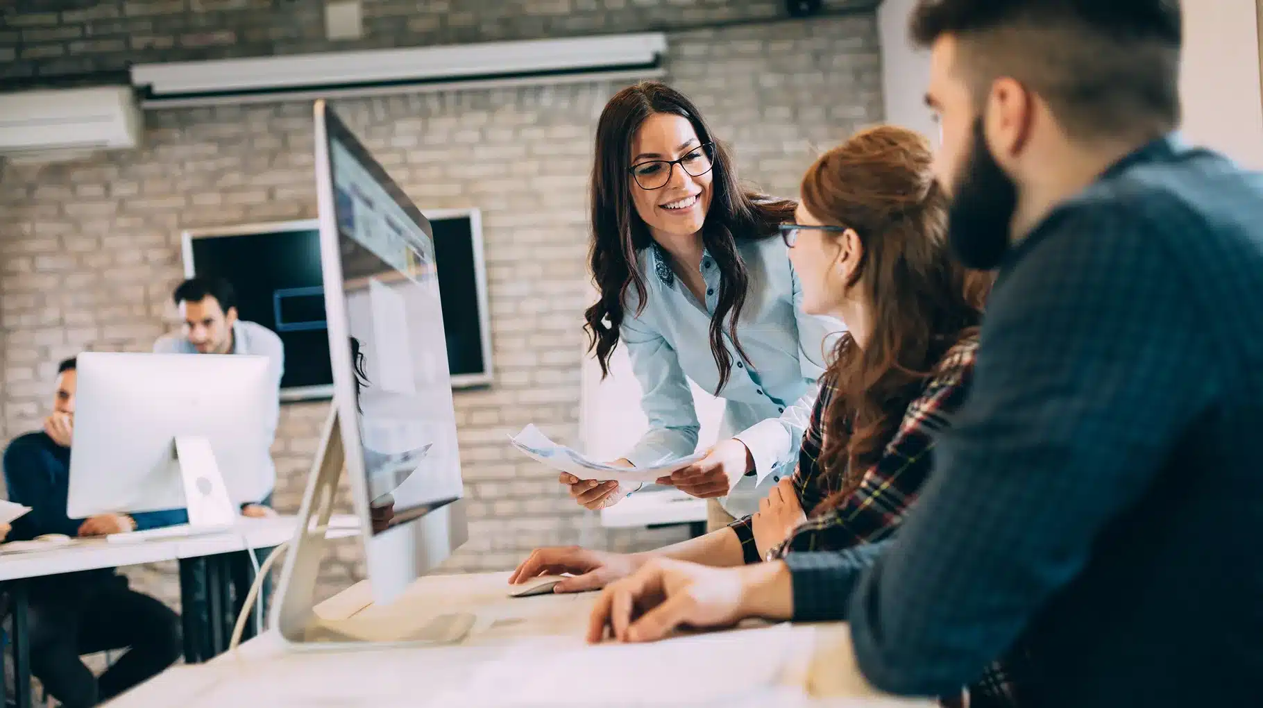 A woman engages in conversation with a man and two women while standing in front of a computer.