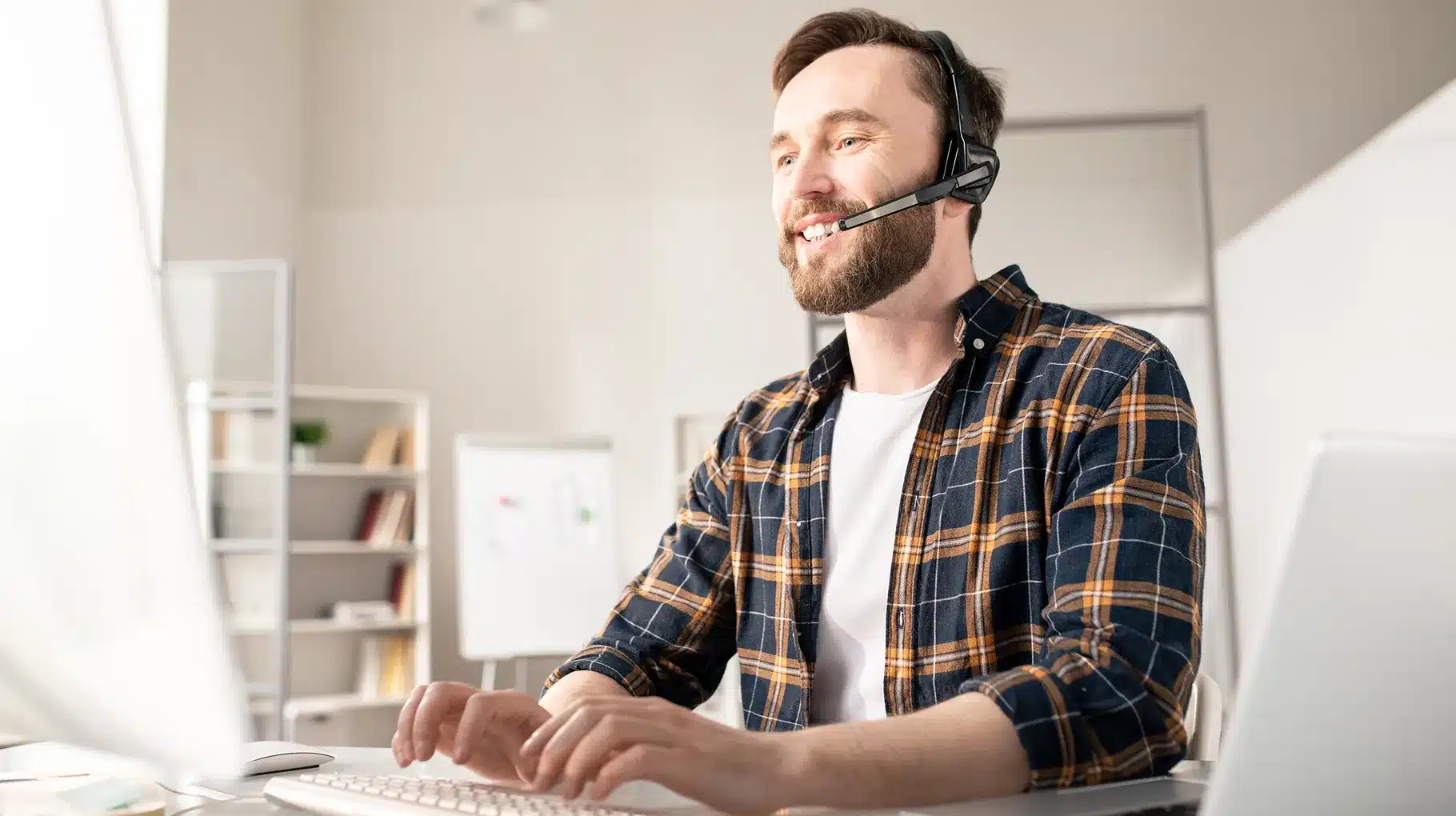 A man with a headset smiles while working on a computer, indicating a positive and engaged work environment.