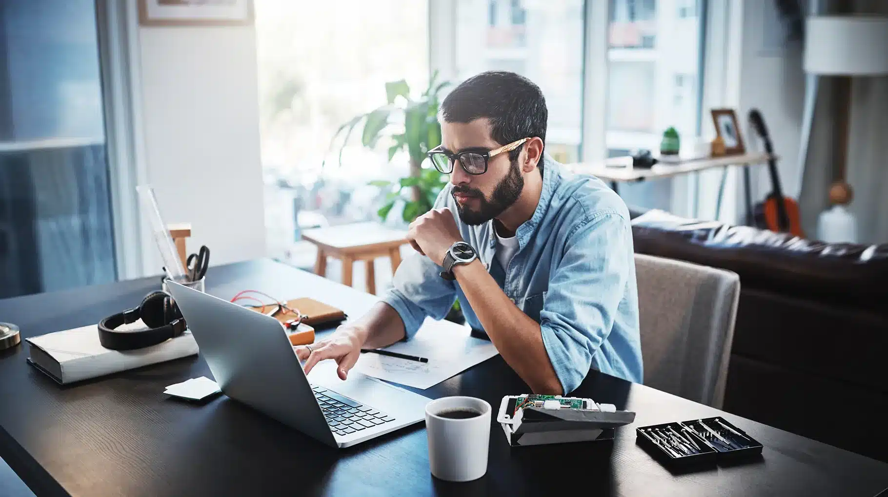 A man wearing glasses sits at a desk, focused on his laptop while working in a well-lit office environment.