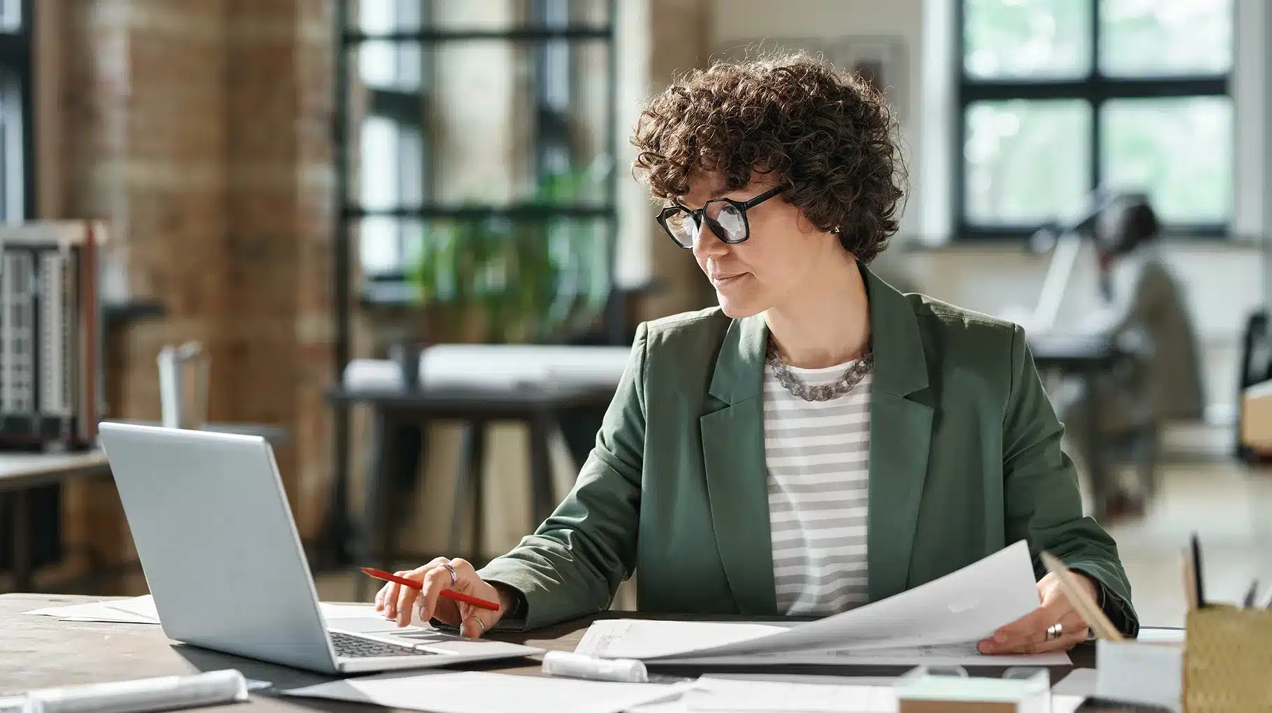 A woman wearing glasses is focused on her laptop, engaged in work at a desk.