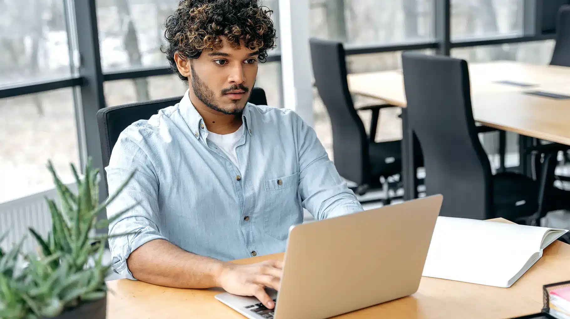 A man sitting at a desk, focused on his laptop, with a notepad and pen beside him.