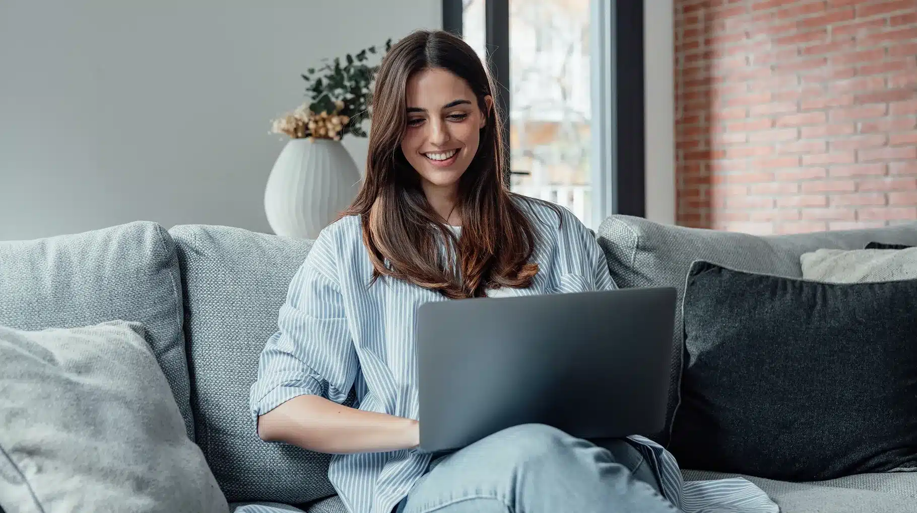 A woman sitting on a couch, focused on her laptop, with a cozy living room setting in the background.