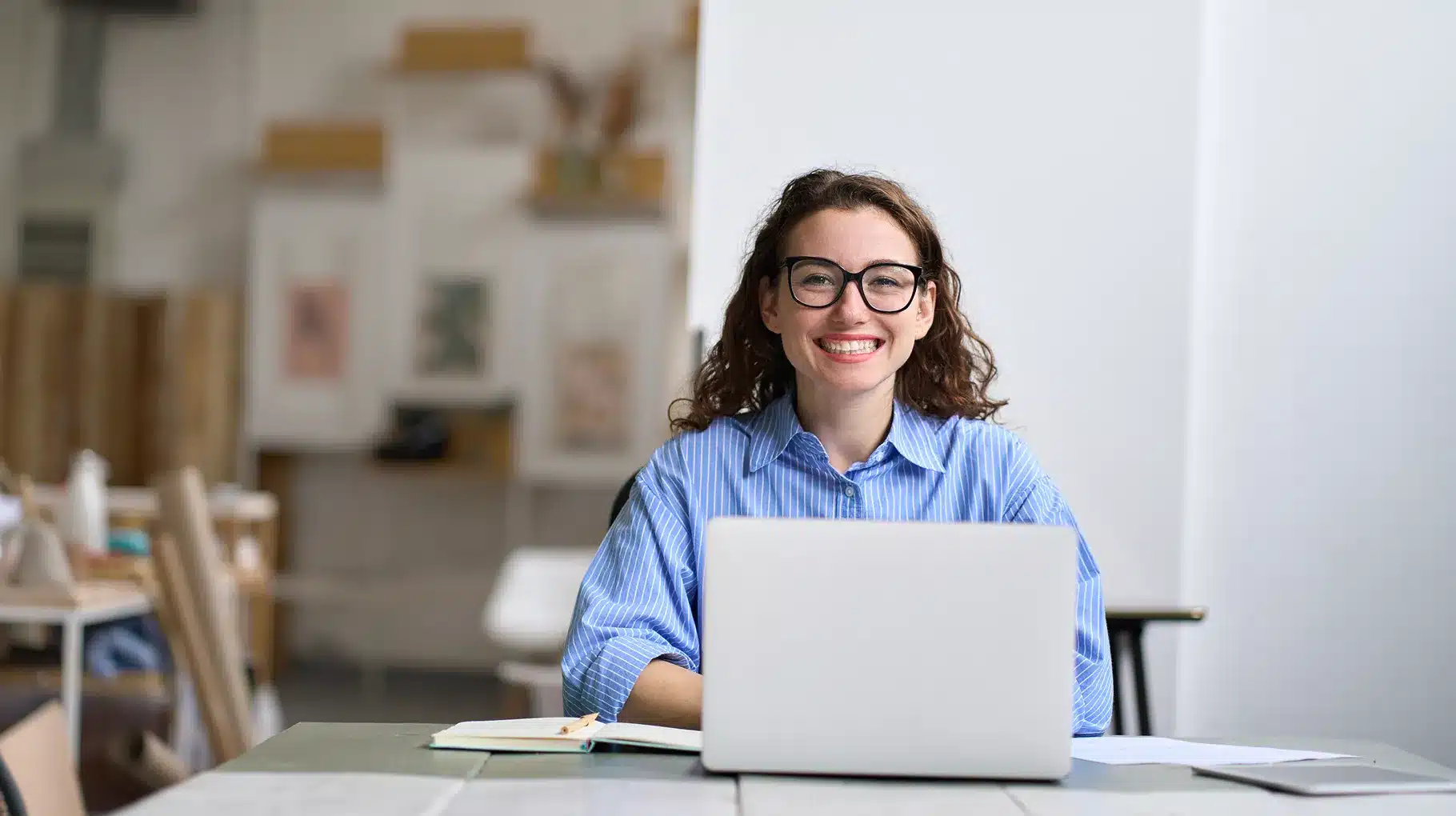 A woman wearing glasses sits at a table, focused on her laptop, with a calm expression in a well-lit environment.