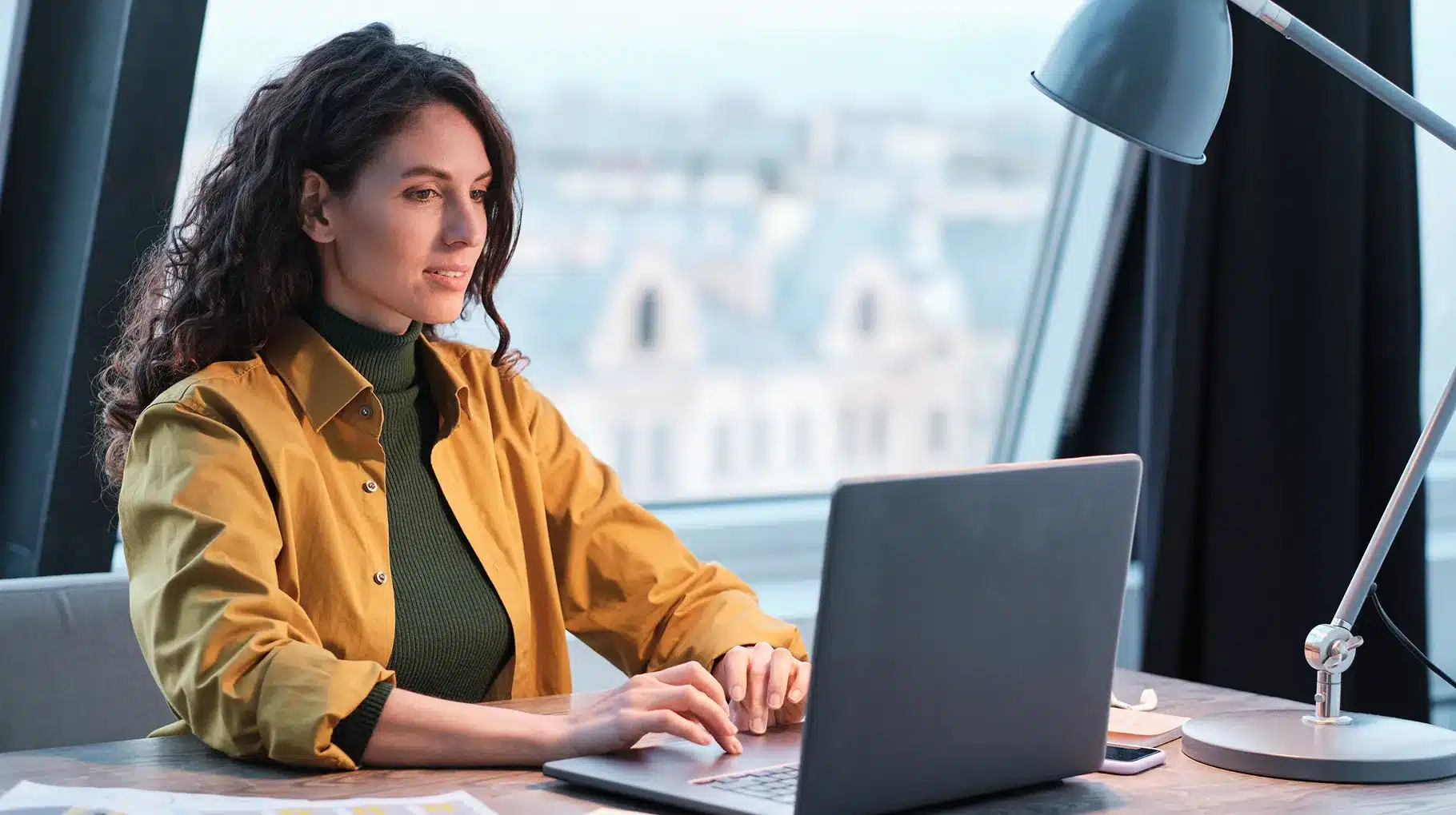 A woman sitting at a desk, focused on her laptop, with a notepad and pen beside her.