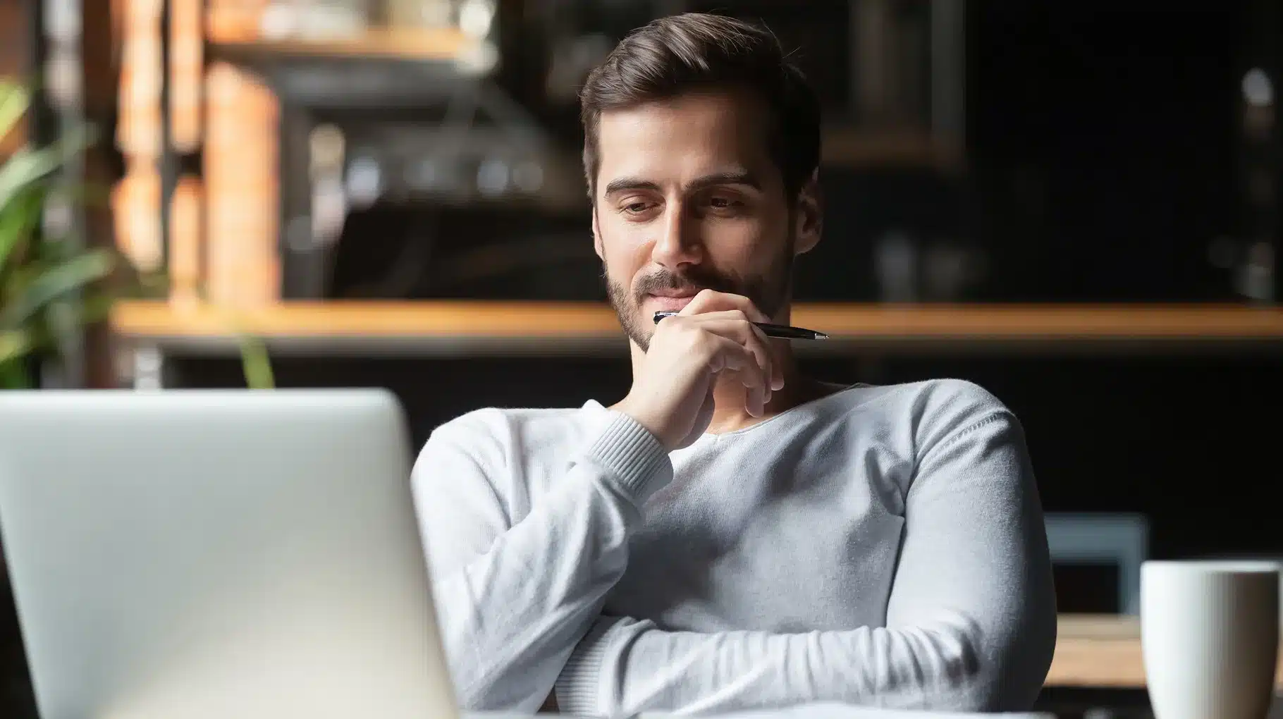 A man sitting at a table, focused on his laptop, with a neutral background.