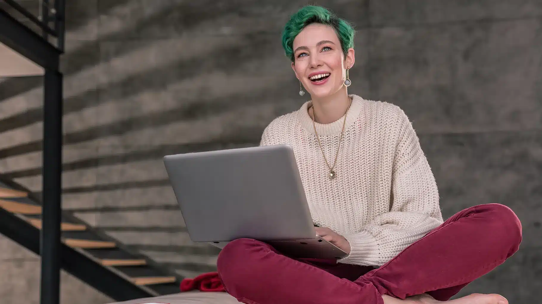 A woman with green hair sits on a bed, using a laptop, surrounded by soft pillows and a cozy blanket.