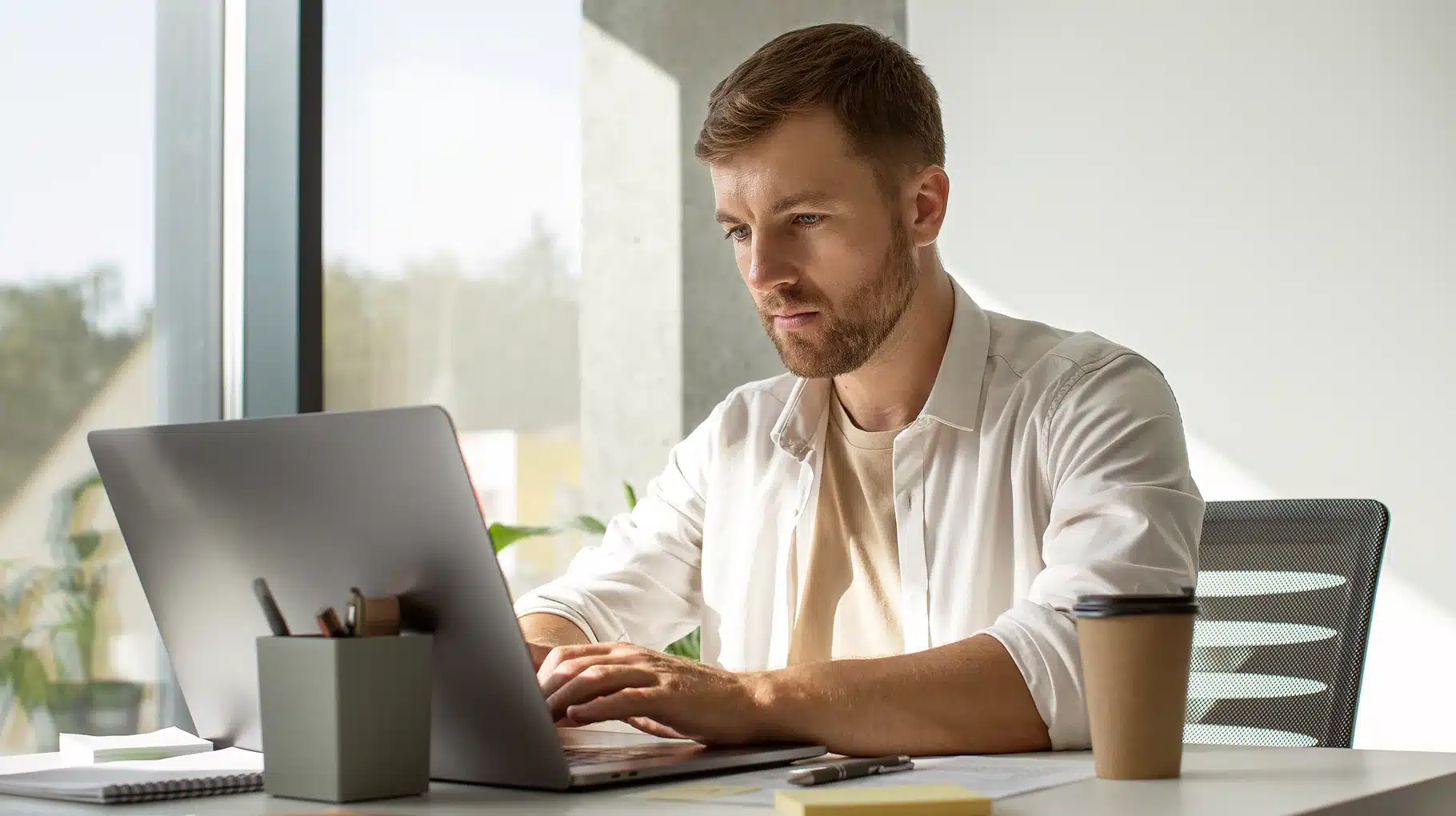 A man sitting at a desk, focused on his laptop, with a notepad and pen beside him.