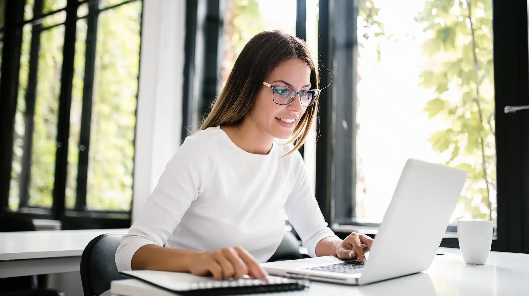 A woman wearing glasses is focused on her laptop, engaged in work at a desk.