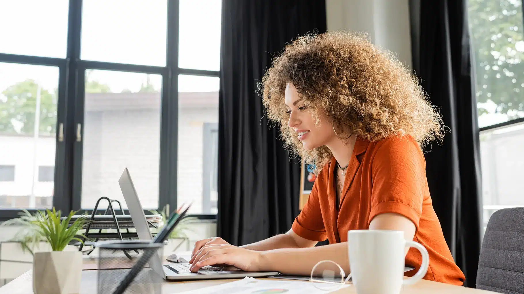 A woman with curly hair sits at a desk, focused on her laptop, surrounded by books and a notepad.
