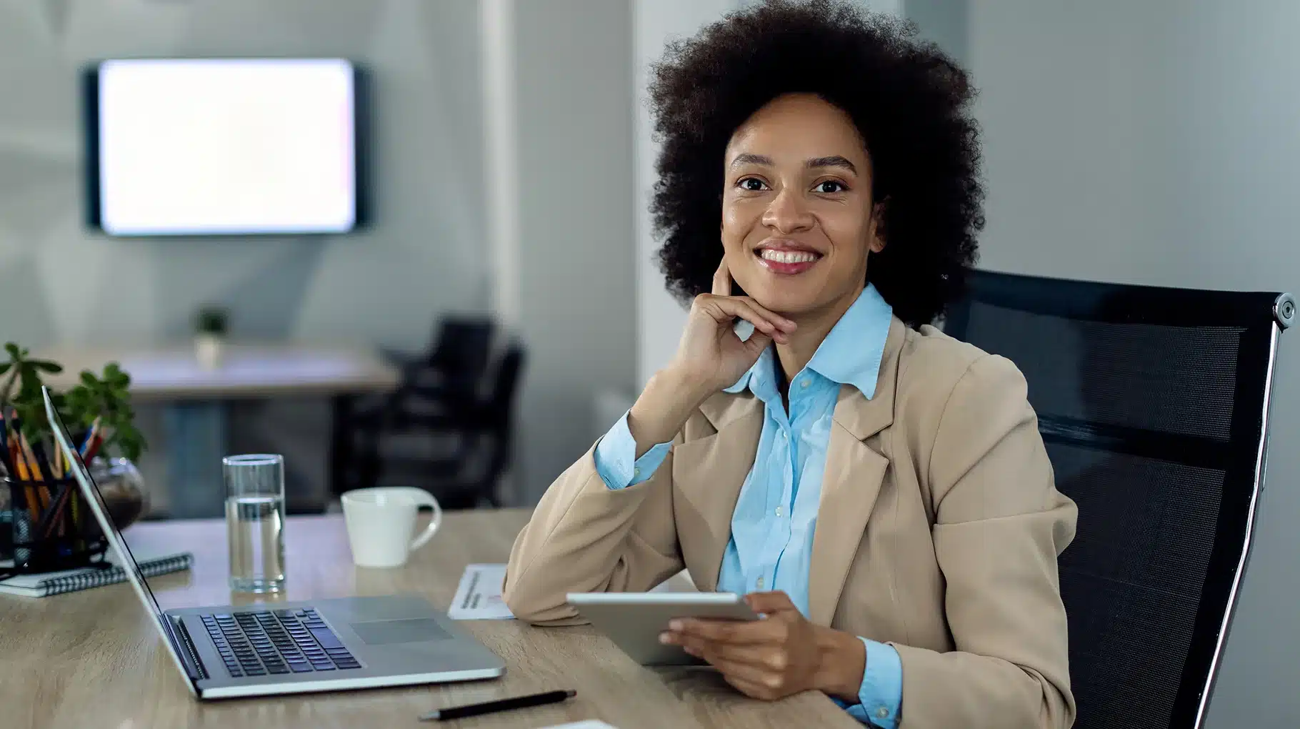 Smiling person in a beige blazer and blue shirt sits at a desk with a tablet. A laptop, glass of water, and coffee cup are nearby, conveying a professional setting.