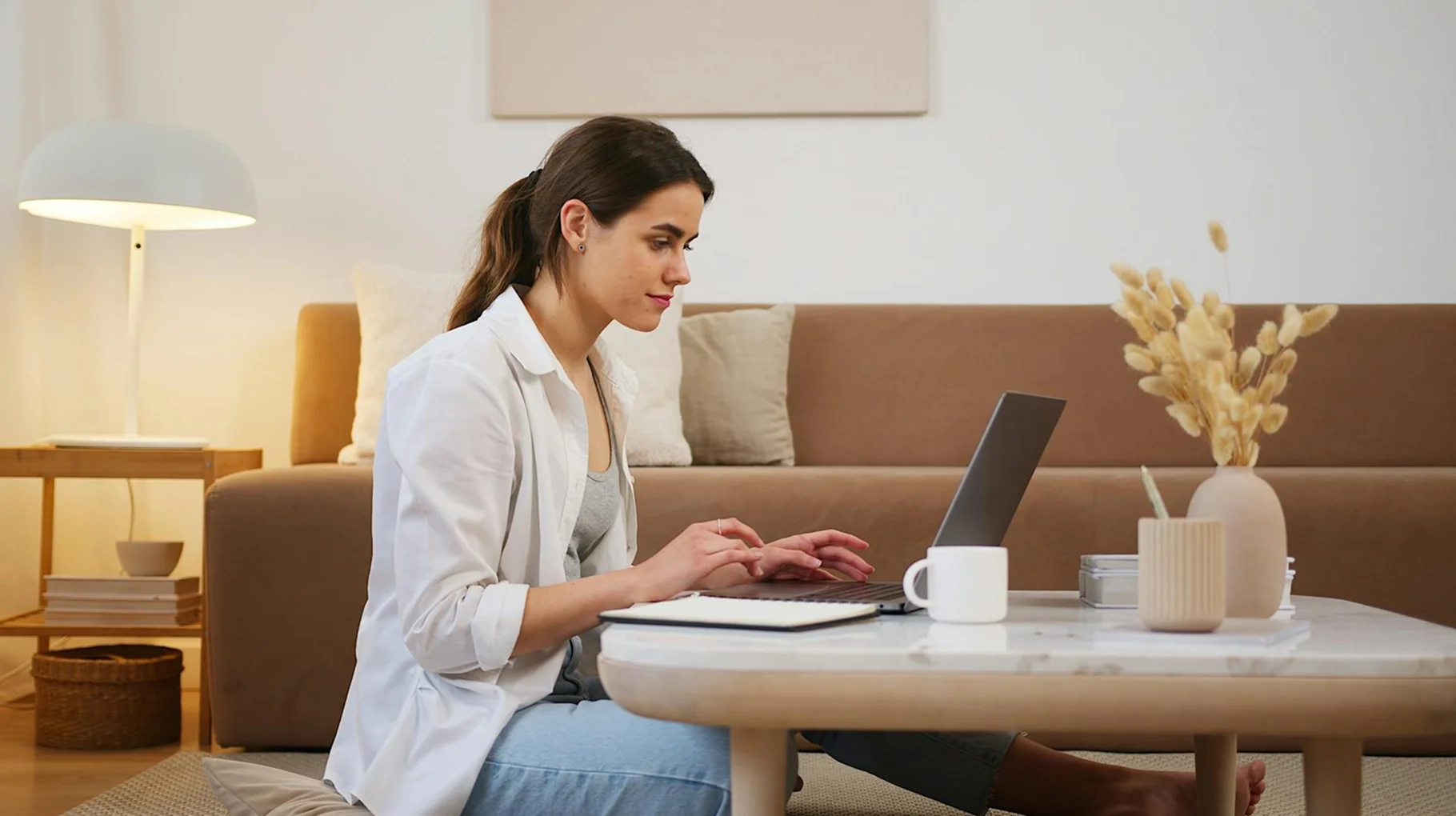 A woman sitting on the floor, focused on her laptop, with a relaxed posture and a cozy environment around her.