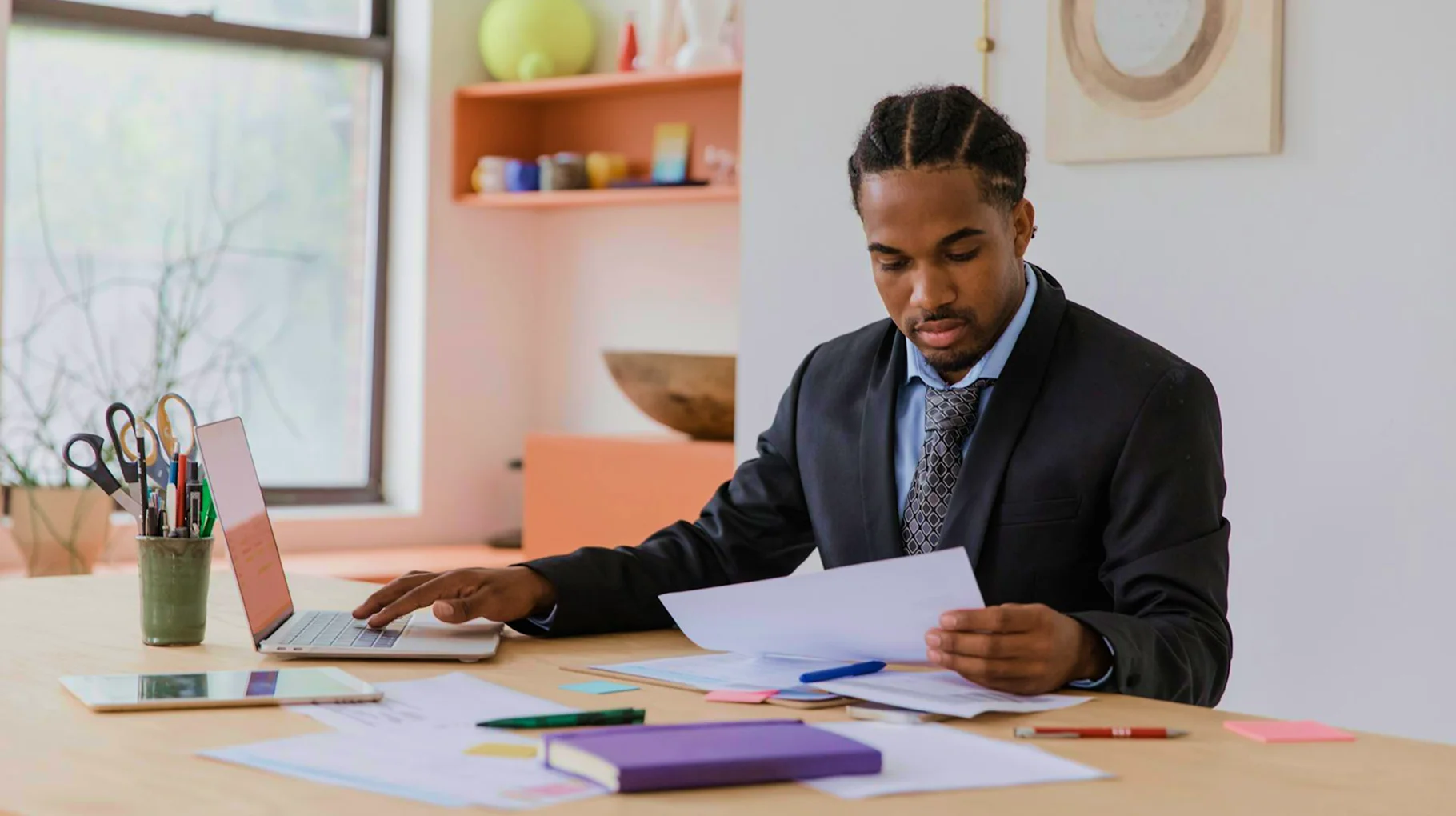 A man in a suit sits at a desk with papers and a laptop, focused on his work.