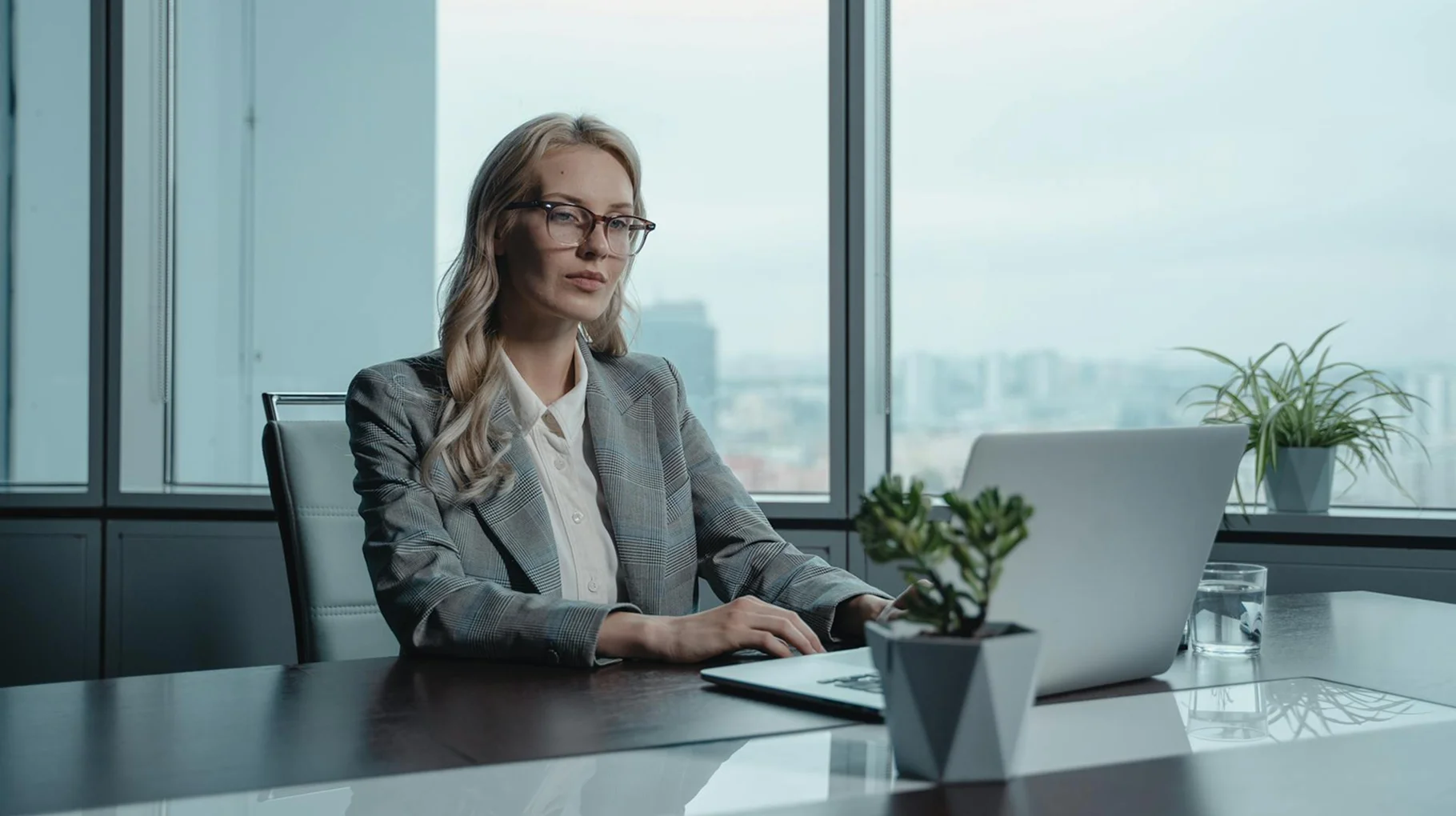 A woman wearing glasses sits at a desk, focused on her laptop while working.
