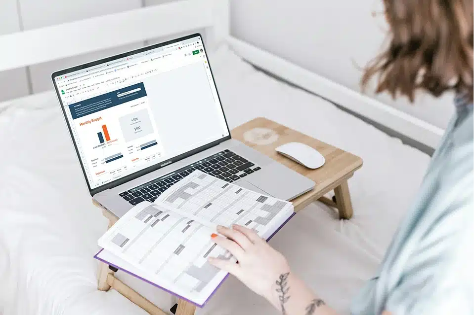A woman sits on a bed, using a laptop while taking notes in a notebook beside her.