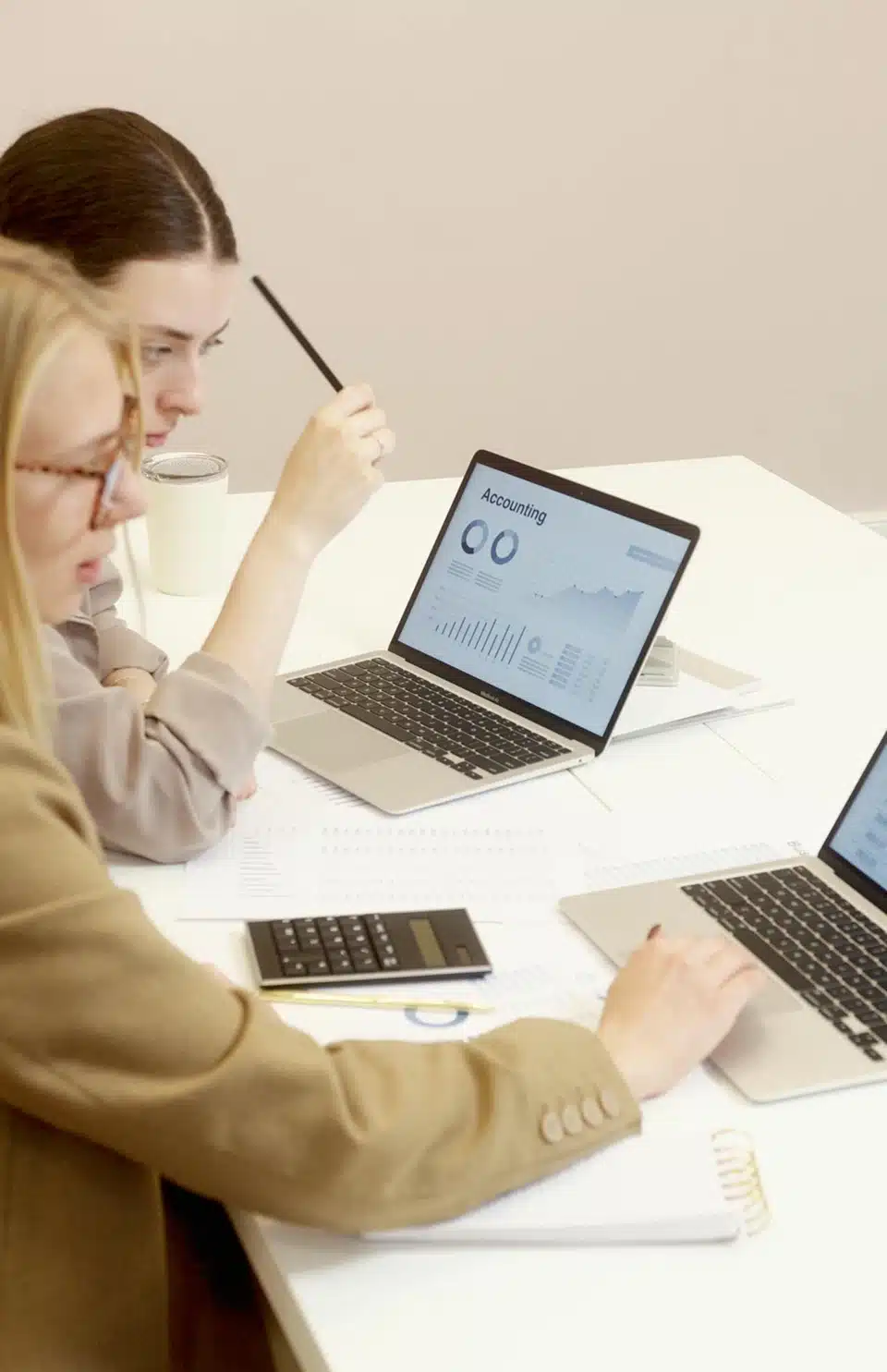Two women sit at a table, each using a laptop, engaged in conversation and collaboration.