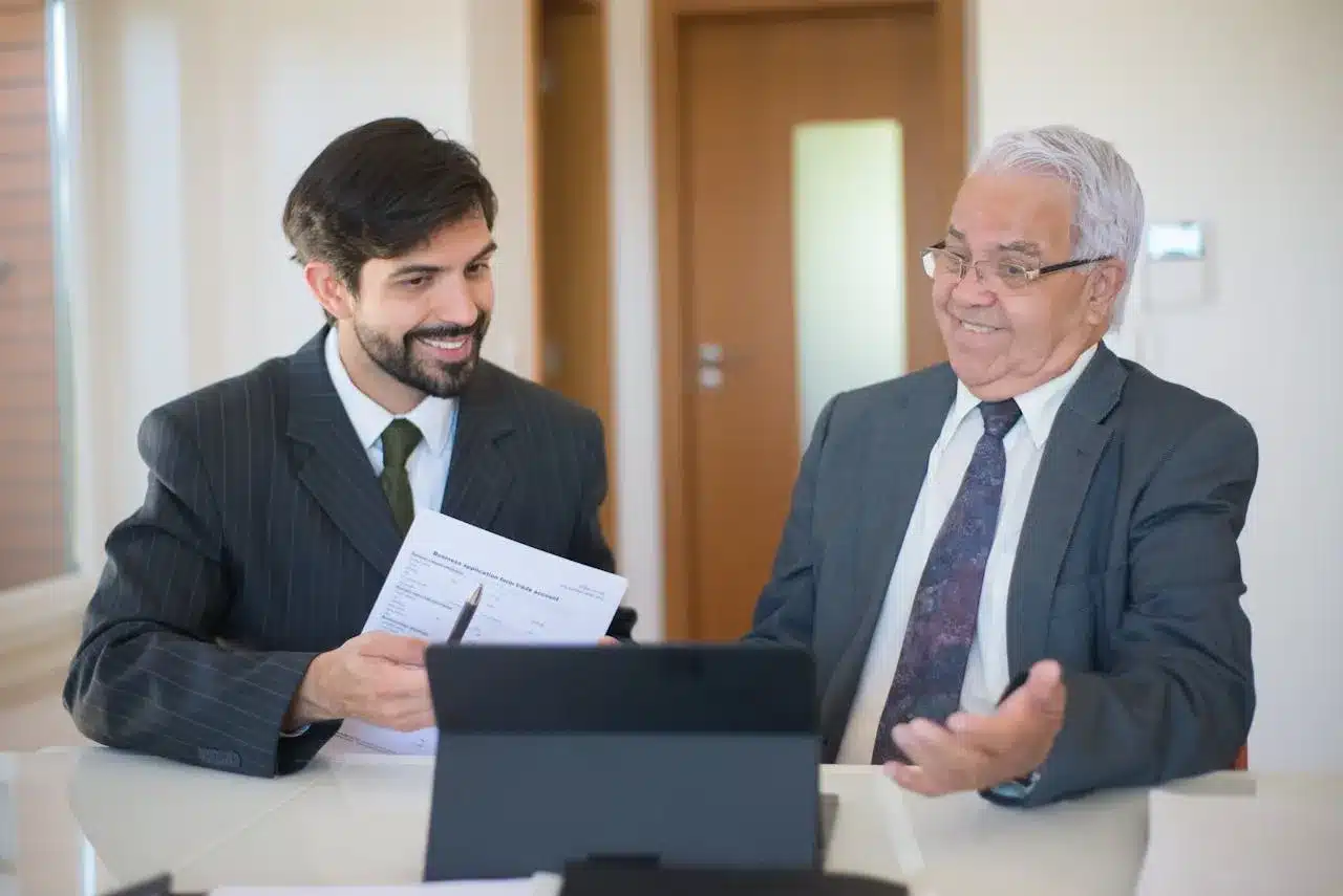 Two men in business suits seated at a table, working together on a laptop.