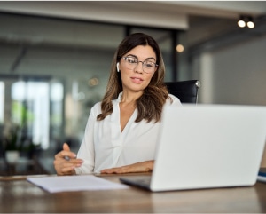 A woman wearing glasses is seated at a desk, focused on her laptop while working.
