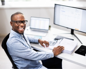 A smiling man wearing glasses sits at a desk, working on a computer, exuding a sense of contentment and focus.