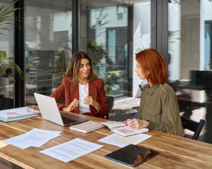 Two women engaged in discussion at a table, with a laptop open between them, indicating collaboration or work.