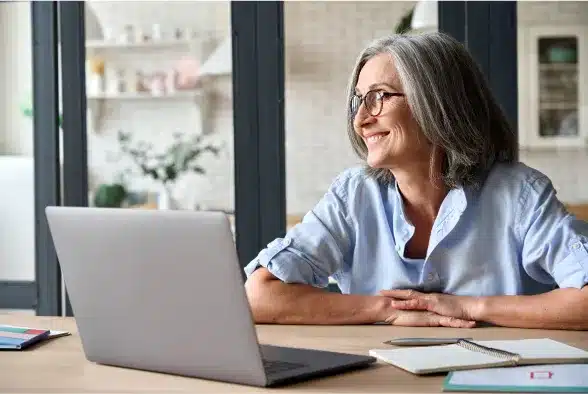 A woman wearing glasses is seated at a table, focused on her laptop.