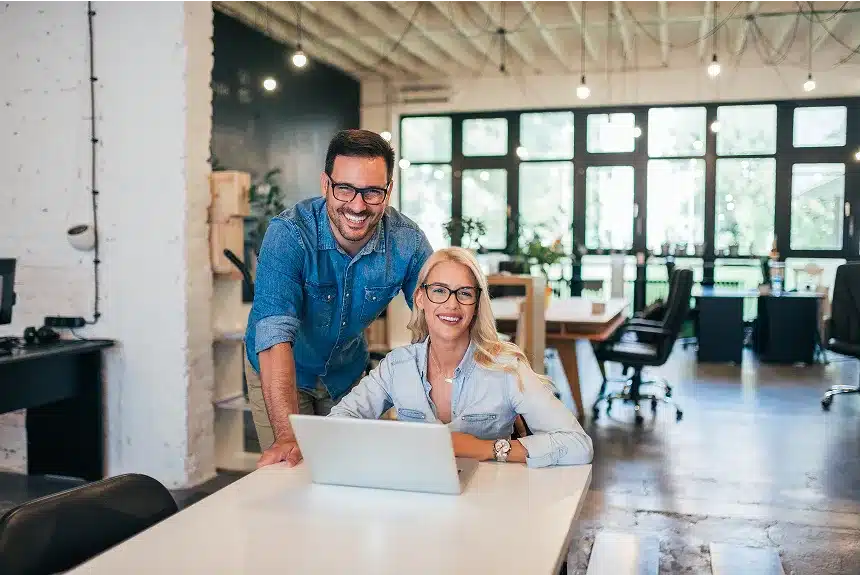 A man and woman smile while sitting at a table, engaged with a laptop in front of them.