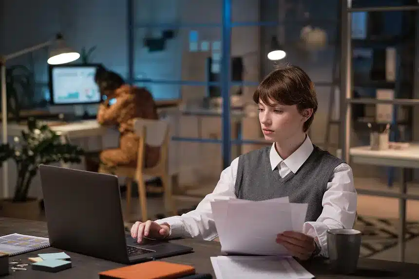 A man wearing glasses sits at a desk, focused on his laptop while working in a well-lit office environment.