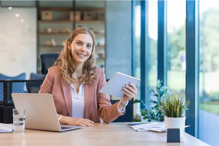A woman sits at a desk, focused on her laptop and tablet, surrounded by a tidy workspace.