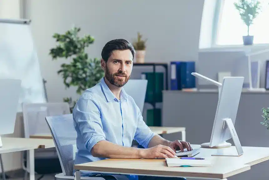A man seated at a desk, focused on his computer screen, with a notepad and pen beside him.