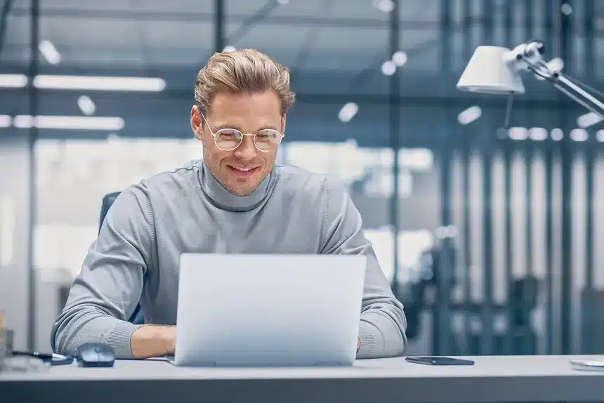 A man wearing glasses is focused on working on his laptop at a desk.