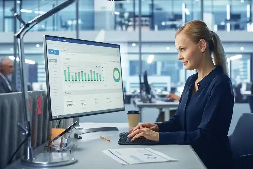 A woman focused on her work, typing on a computer in front of a large monitor in a well-lit office environment.