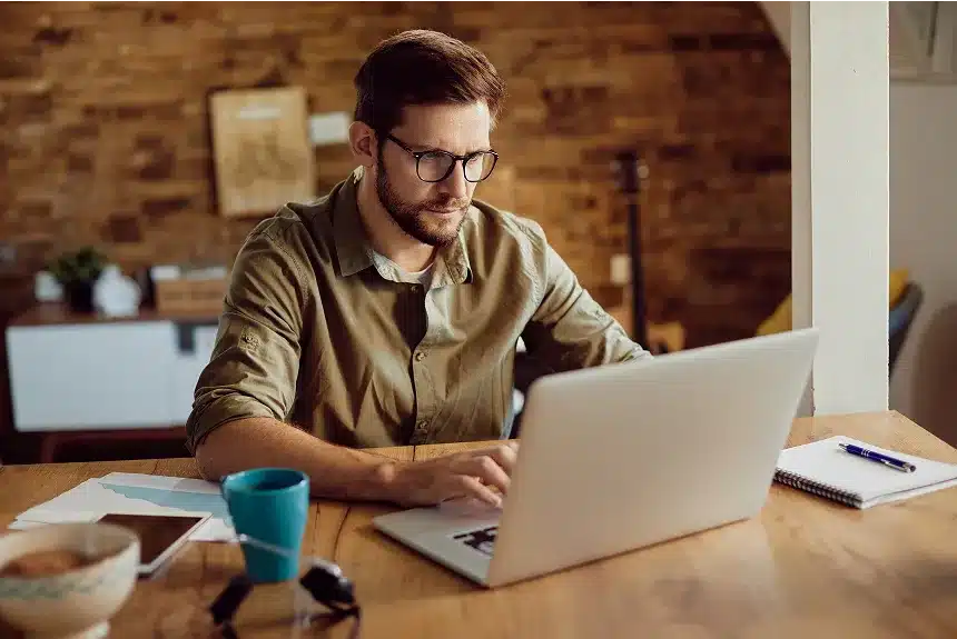 A man wearing glasses sits at a table, focused on his laptop while working.