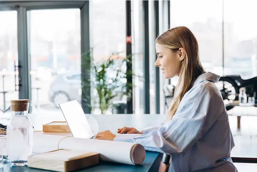 A woman sitting at a table, focused on her laptop, with a calm expression and a cozy indoor setting.