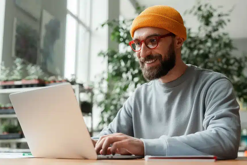 A bearded man wearing glasses is focused on working on his laptop at a desk.