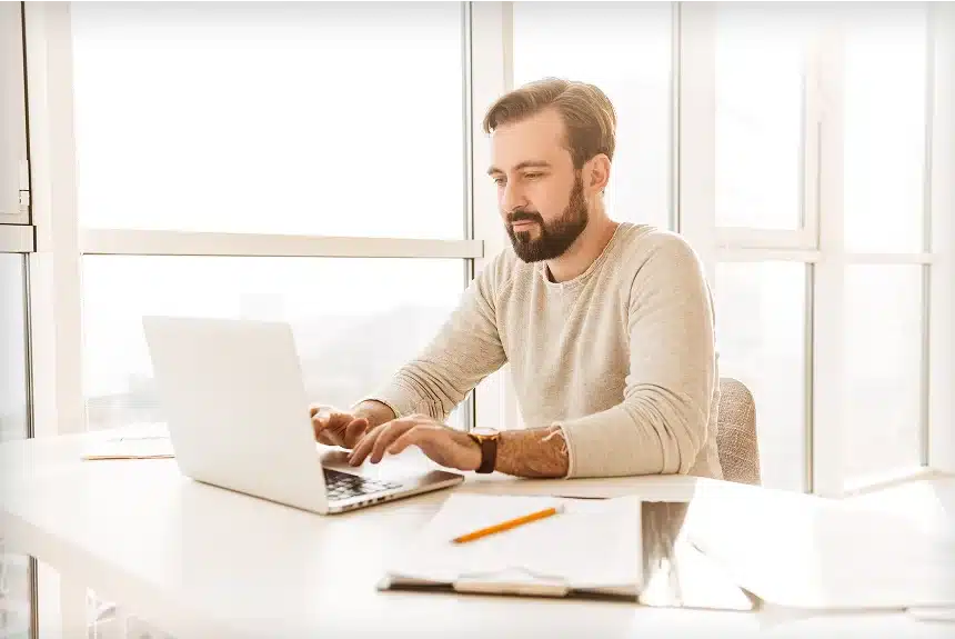 A bearded man sitting at a desk, focused on his laptop while working.