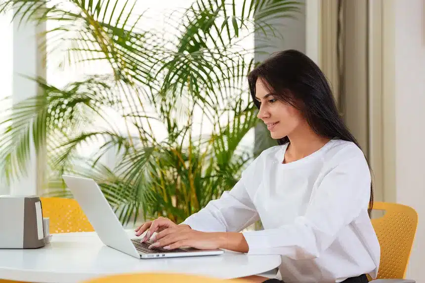 A woman seated at a table, focused on her laptop, with a calm expression in a well-lit room.