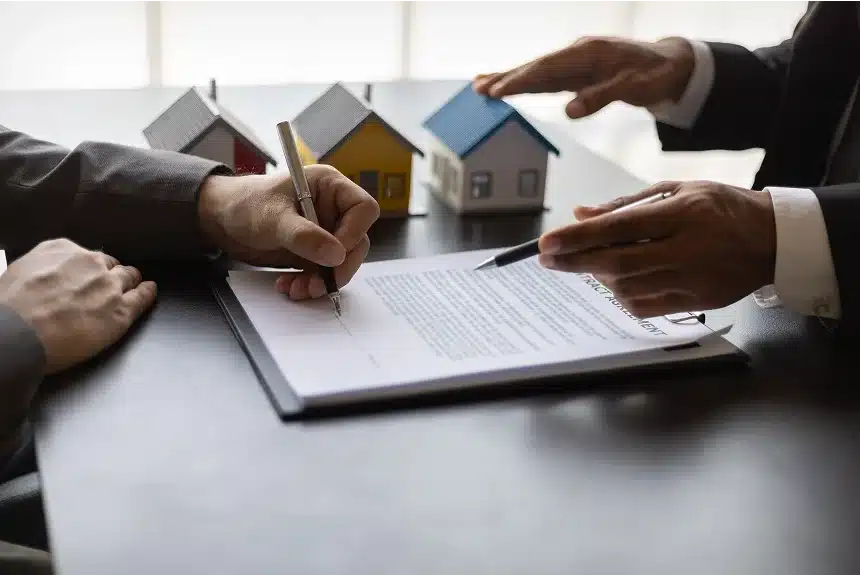 Two people discussing a contract, with one signing a document. Three small house models on the table suggest a real estate transaction. Professional tone.