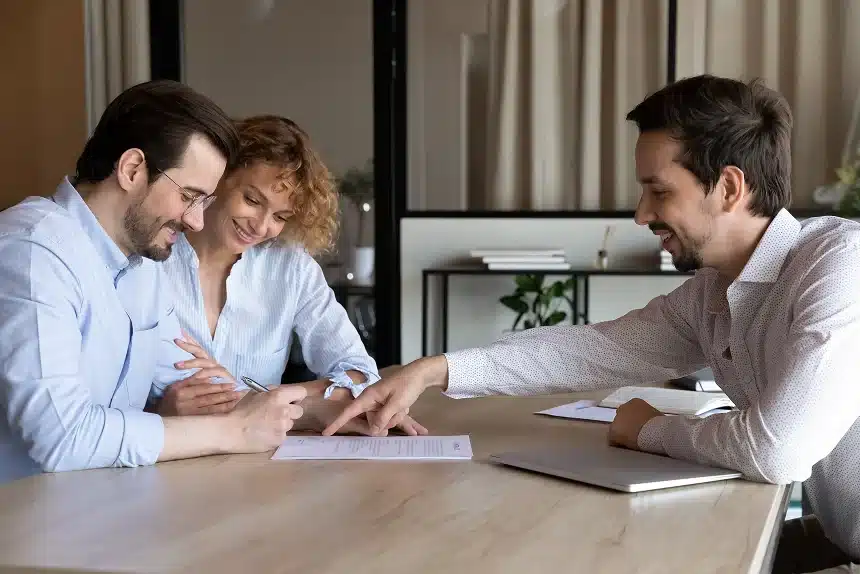 A couple sits at a table signing a document while a smiling advisor points at the paper. The setting is professional and collaborative.