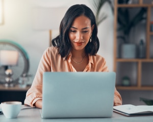A woman seated at a desk, focused on her laptop, surrounded by a tidy workspace and natural light.