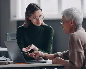Two women engaged in conversation while using a laptop in a professional office setting.