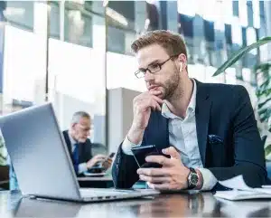 A man in a suit sits at a desk, focused on his phone while engaged in work-related tasks.