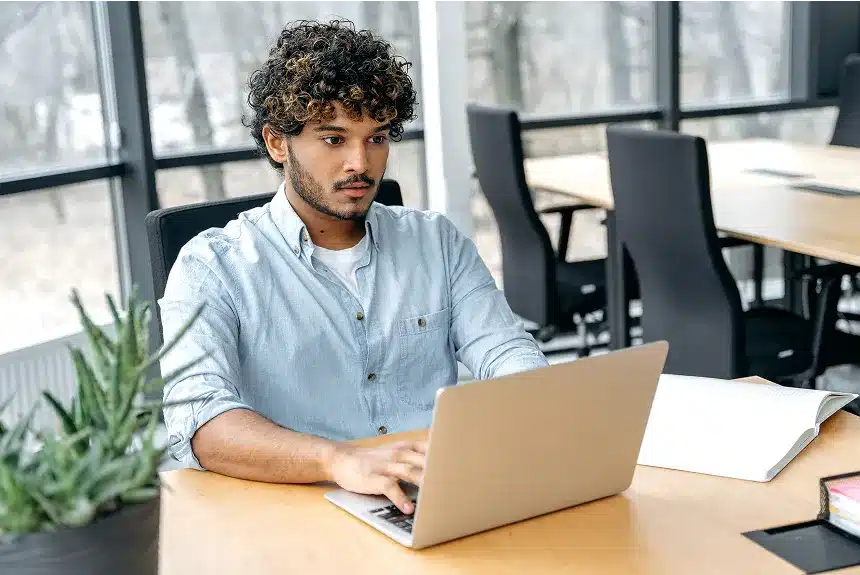A man sitting at a desk, focused on his laptop, with a notepad and pen beside him.