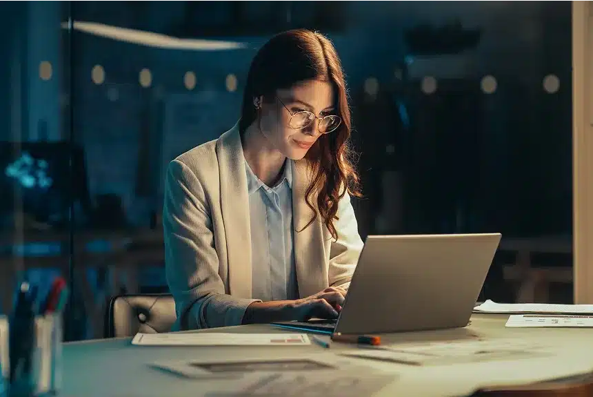 A woman in a business suit focused on her laptop, engaged in work at a modern office setting.