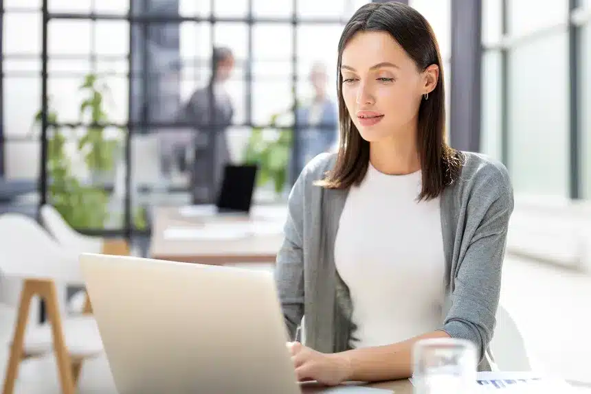 A woman sits at a table, focused on her laptop, with a calm expression in a well-lit indoor setting.