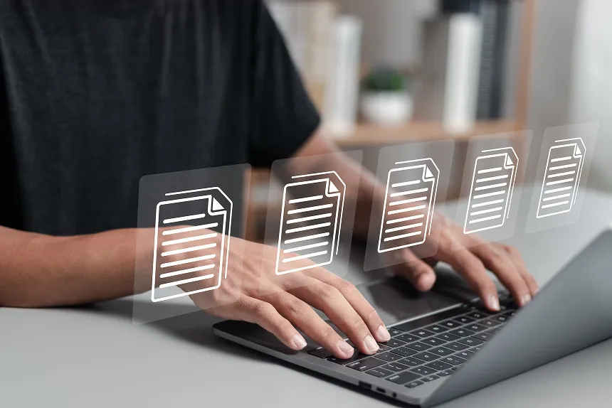 Man typing on a laptop with paper documents displayed on the screen, focused on his work in a well-lit environment.