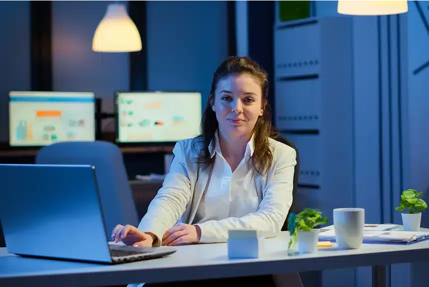 A woman sitting at a desk, focused on her laptop, with a notepad and pen beside her.